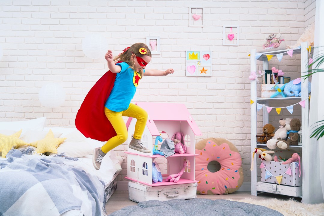 attractive cute little girl jumping from bed to fly, she plays superhero with cloak and mask at home in kids bedroom.concept of bravery and courage in face of small child.feeling of freedom in flight